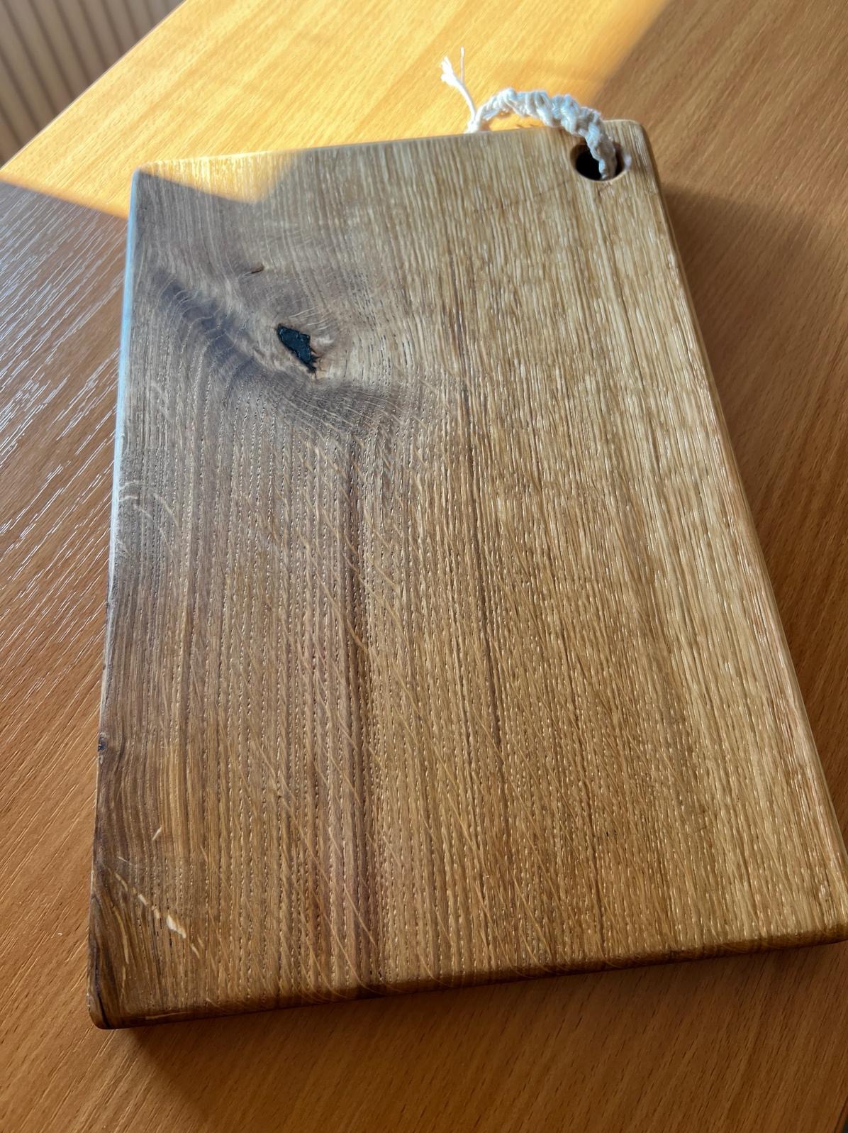 Dębowa deska do krojenia z wyraźnym usłojeniem i małym sękiem, zawieszona na jutowym sznurku, leży na drewnianym blacie w ciepłym świetle.