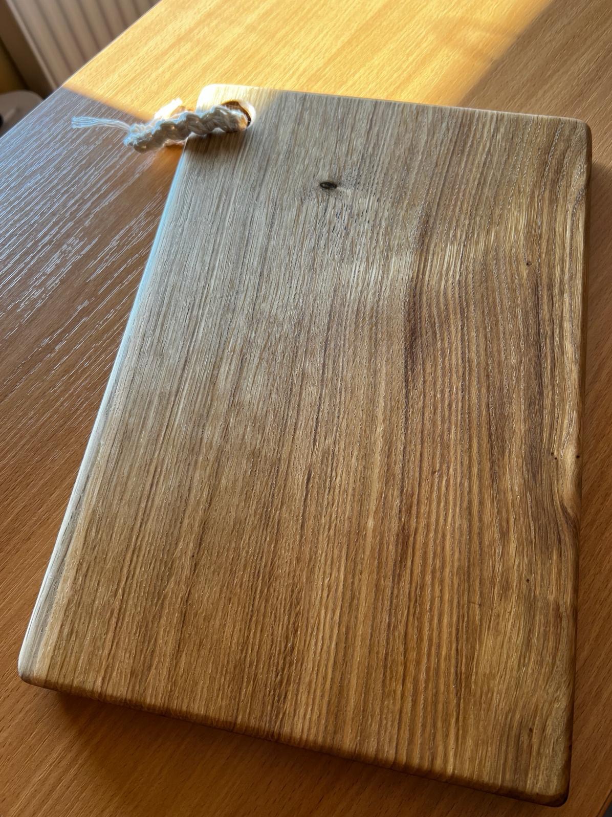 Dębowa deska do krojenia z zawieszką ze sznurka, leżąca na drewnianym blacie w jasnym świetle słonecznym, ukazująca naturalne usłojenie drewna i wykończenie olejem.