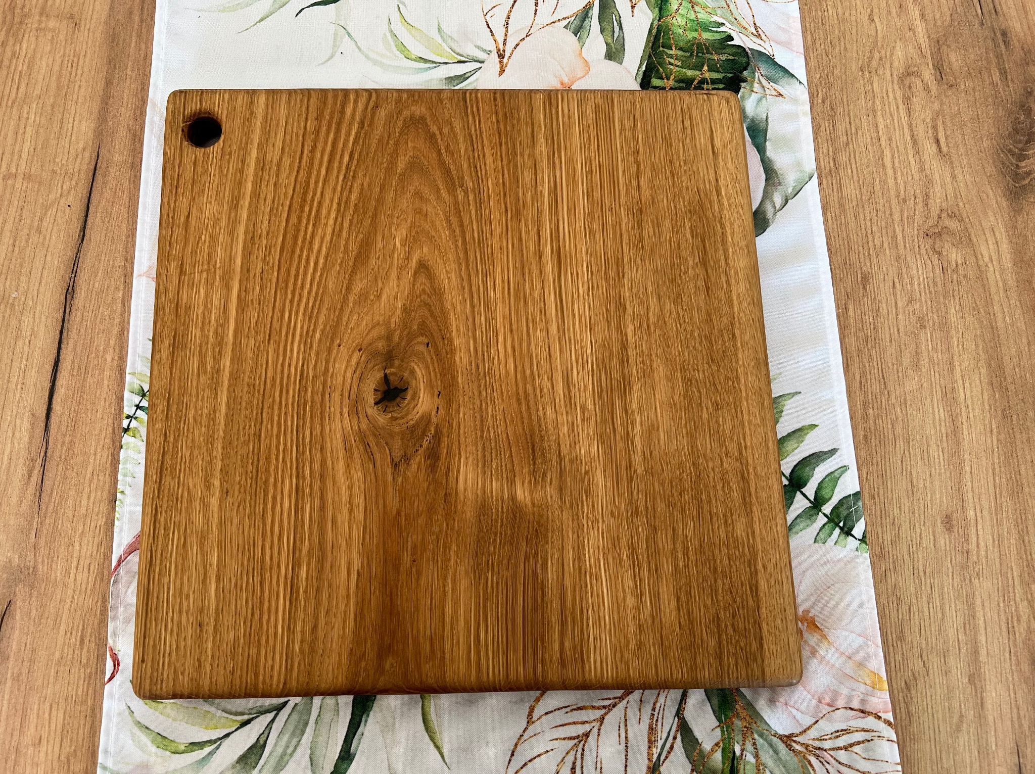 Kwadratowa deska do krojenia z dębowego drewna, z widocznym usłojeniem i otworem w lewym górnym rogu, ułożona na dekoracyjnej podkładce z motywem roślinnym na drewnianym blacie.