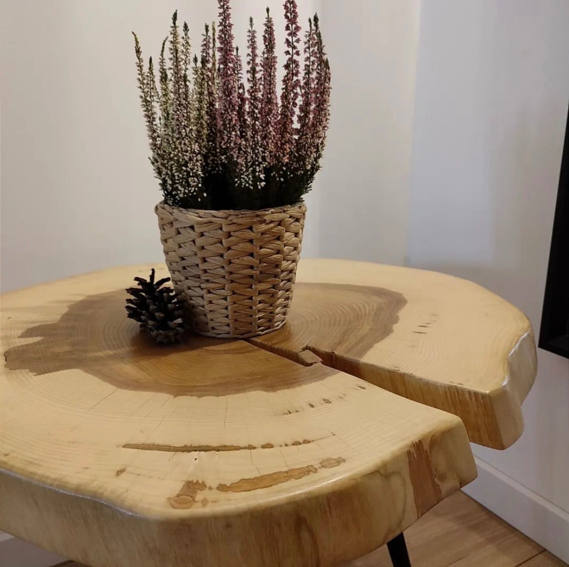 Stolik kawowy z dwóch plastrów drewna jesionowego, wykończony półmatowym olejowoskiem OSMO, z dekoracją w postaci wrzosu w plecionym koszyczku i szyszki.