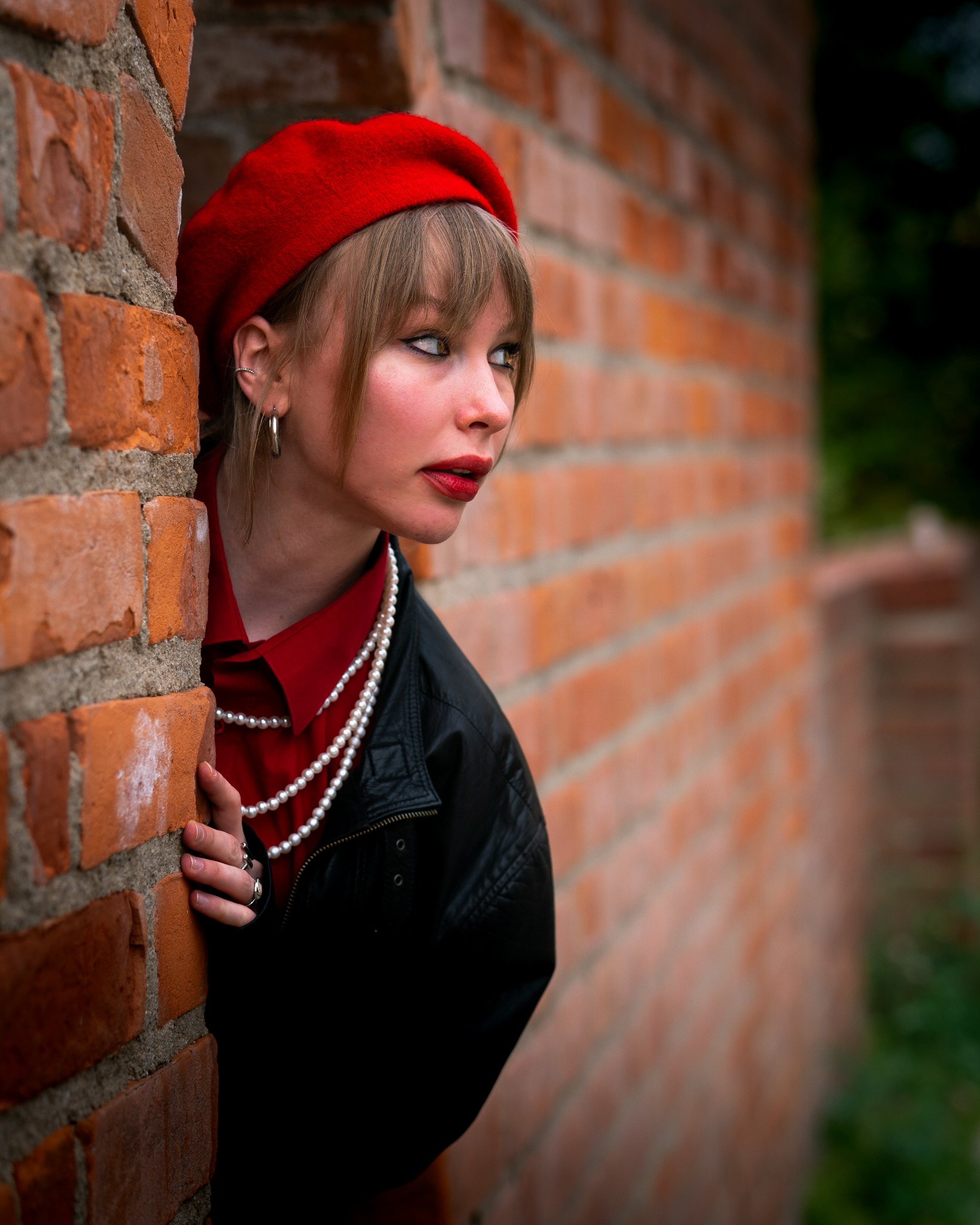 Portret kobiety w czerwonym berecie, opierającej się o ceglaną ścianę, z delikatnym makijażem i perłami na szyi, spoglądającej w bok.