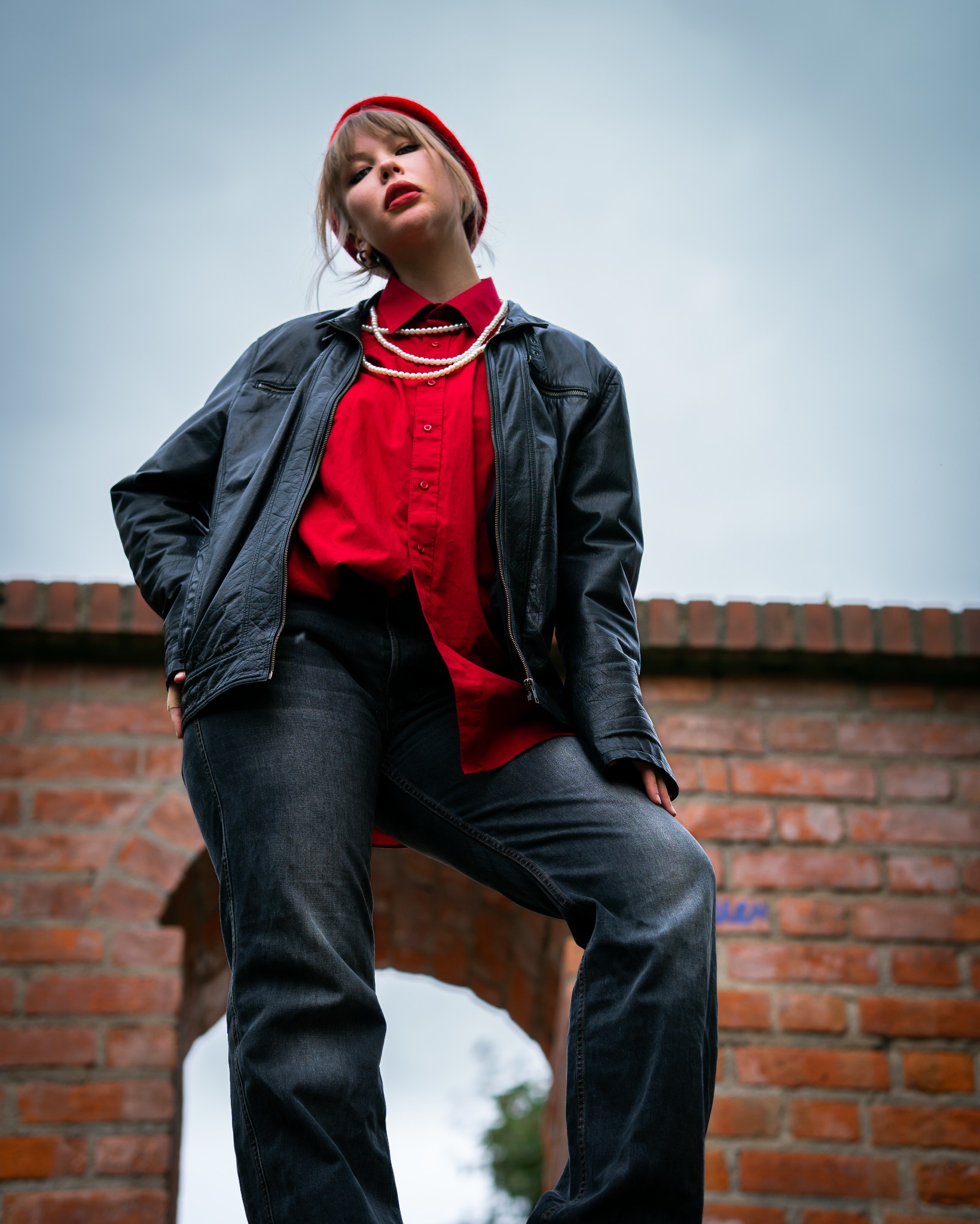 Kobieta w czerwonym berecie, czerwonej koszuli i czarnej skórzanej kurtce pozuje na tle ceglanego łuku, ujęcie z dołu.
