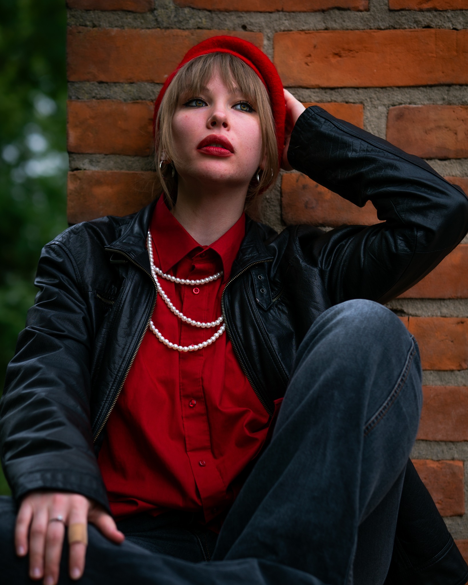 Portret kobiety w czerwonym berecie, czarnej skórzanej kurtce i perłowym naszyjniku, opierającej się o ceglaną ścianę, zamyślona poza.