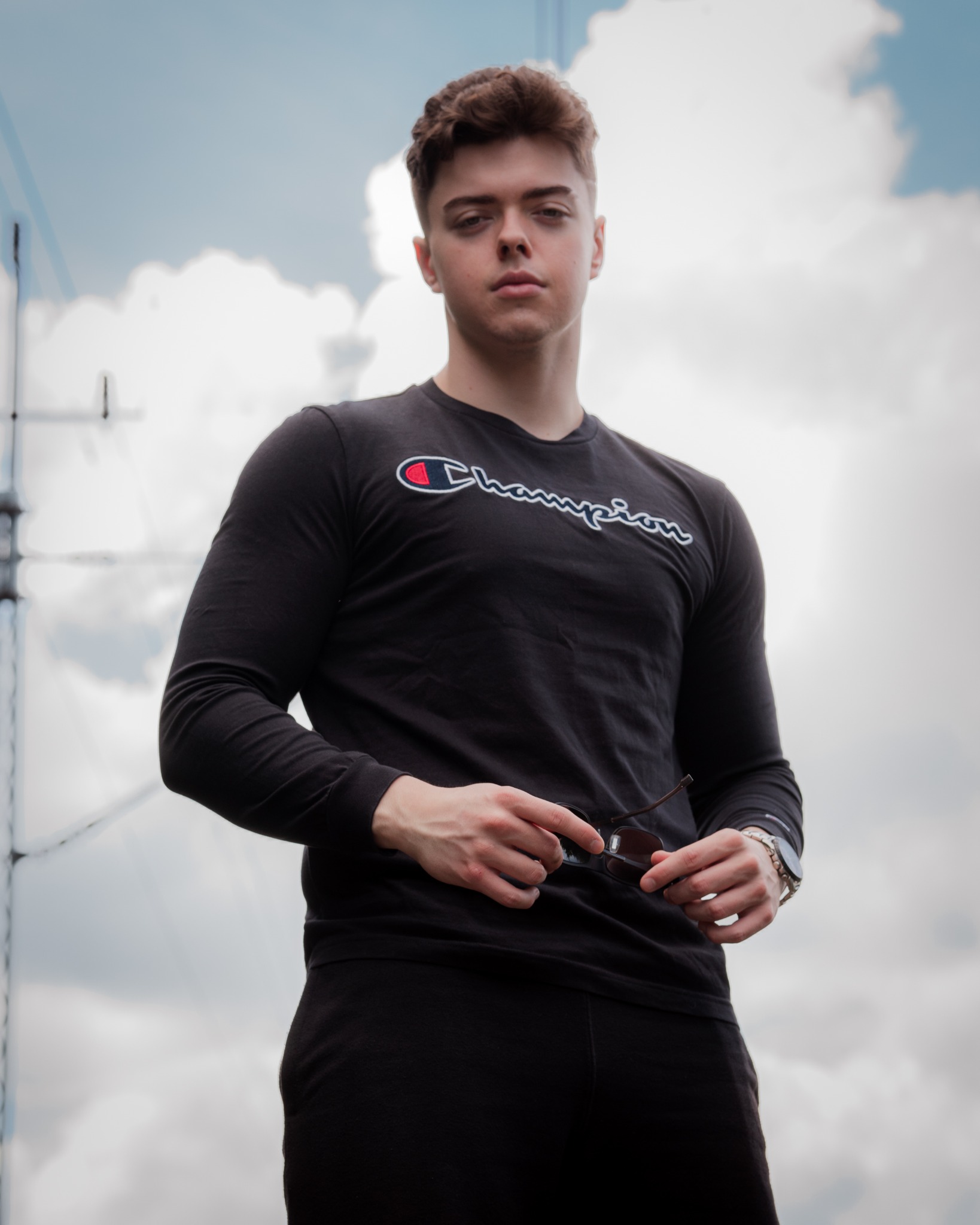 Portret młodego mężczyzny w czarnym stroju sportowym Champion, trzymającego okulary przeciwsłoneczne, na tle pochmurnego nieba i elementów konstrukcyjnych.