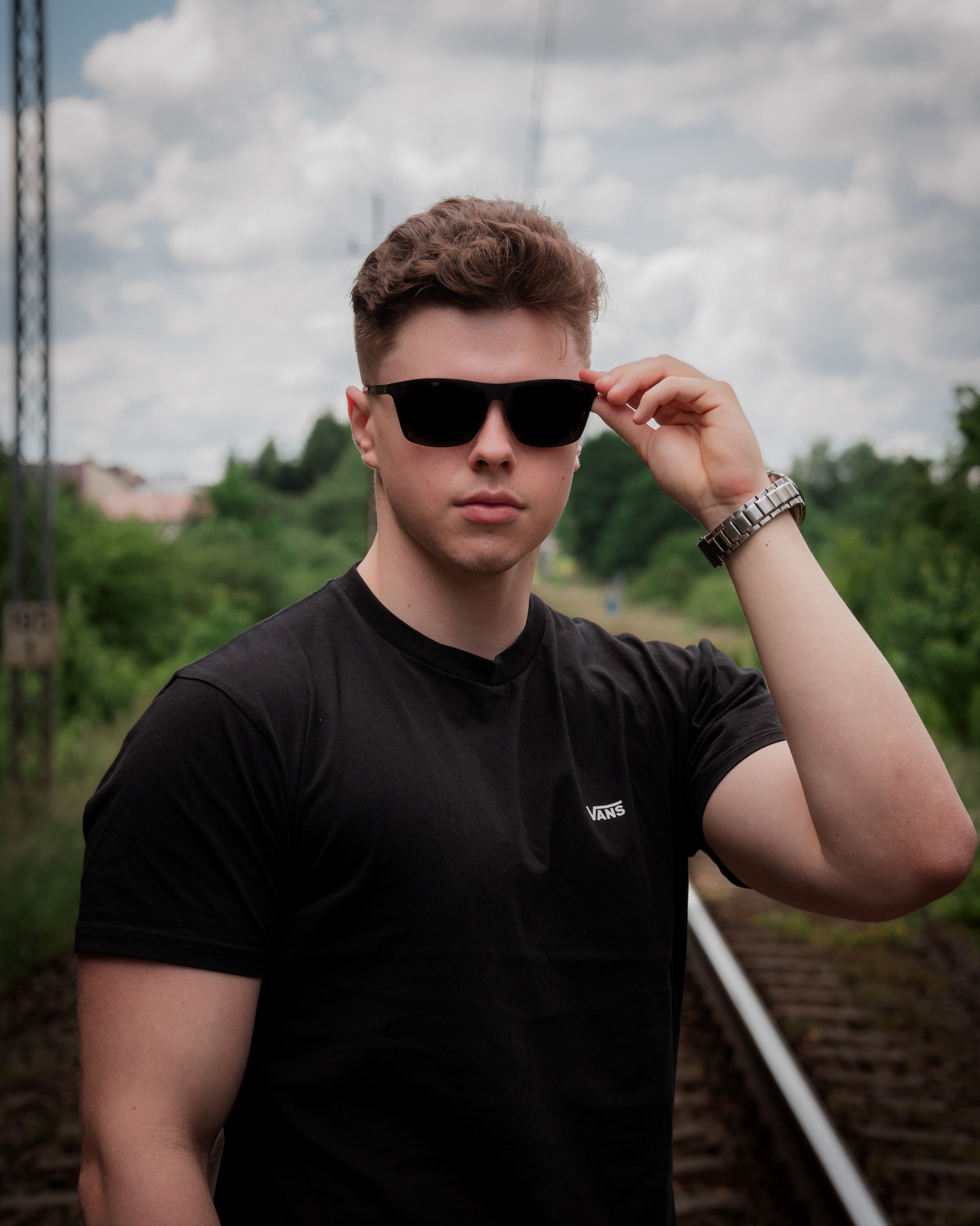 Młody mężczyzna w czarnej koszulce Vans i okularach przeciwsłonecznych, stojący na torach kolejowych, z ręką unoszącą okulary, prezentujący srebrny zegarek.