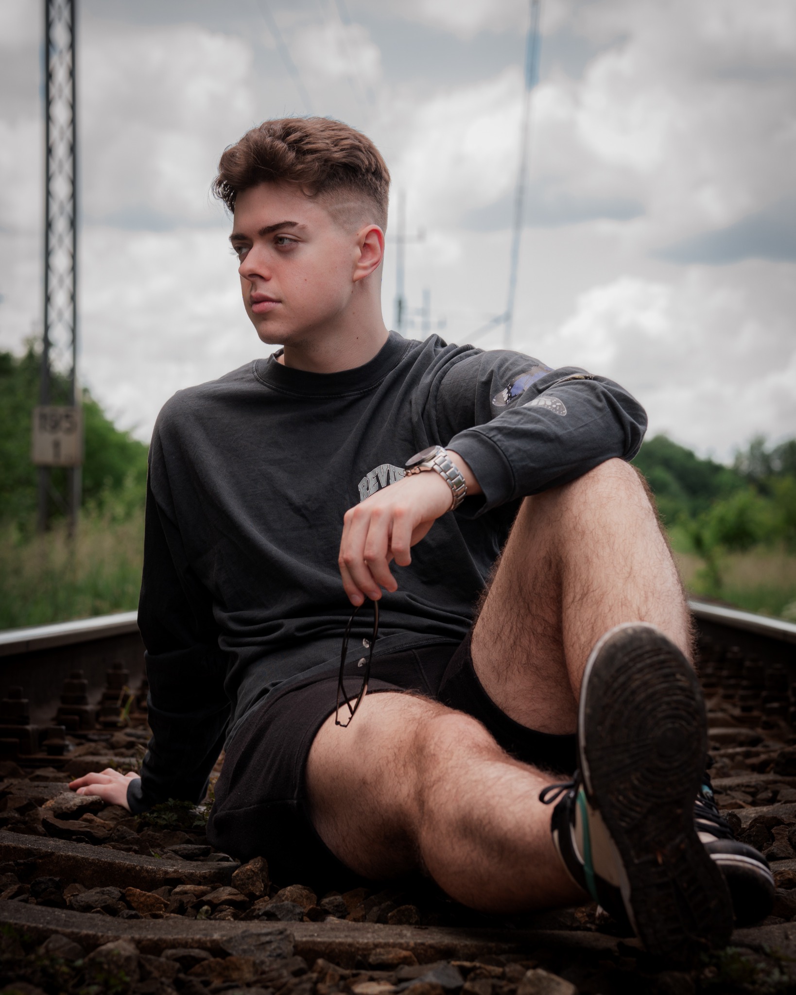Młody mężczyzna z modną fryzurą siedzi na torach kolejowych, ubrany w czarną bluzę i szorty, trzymając okulary w dłoni, z zamyślonym wyrazem twarzy i stalowym zegarkiem na nadgarstku.