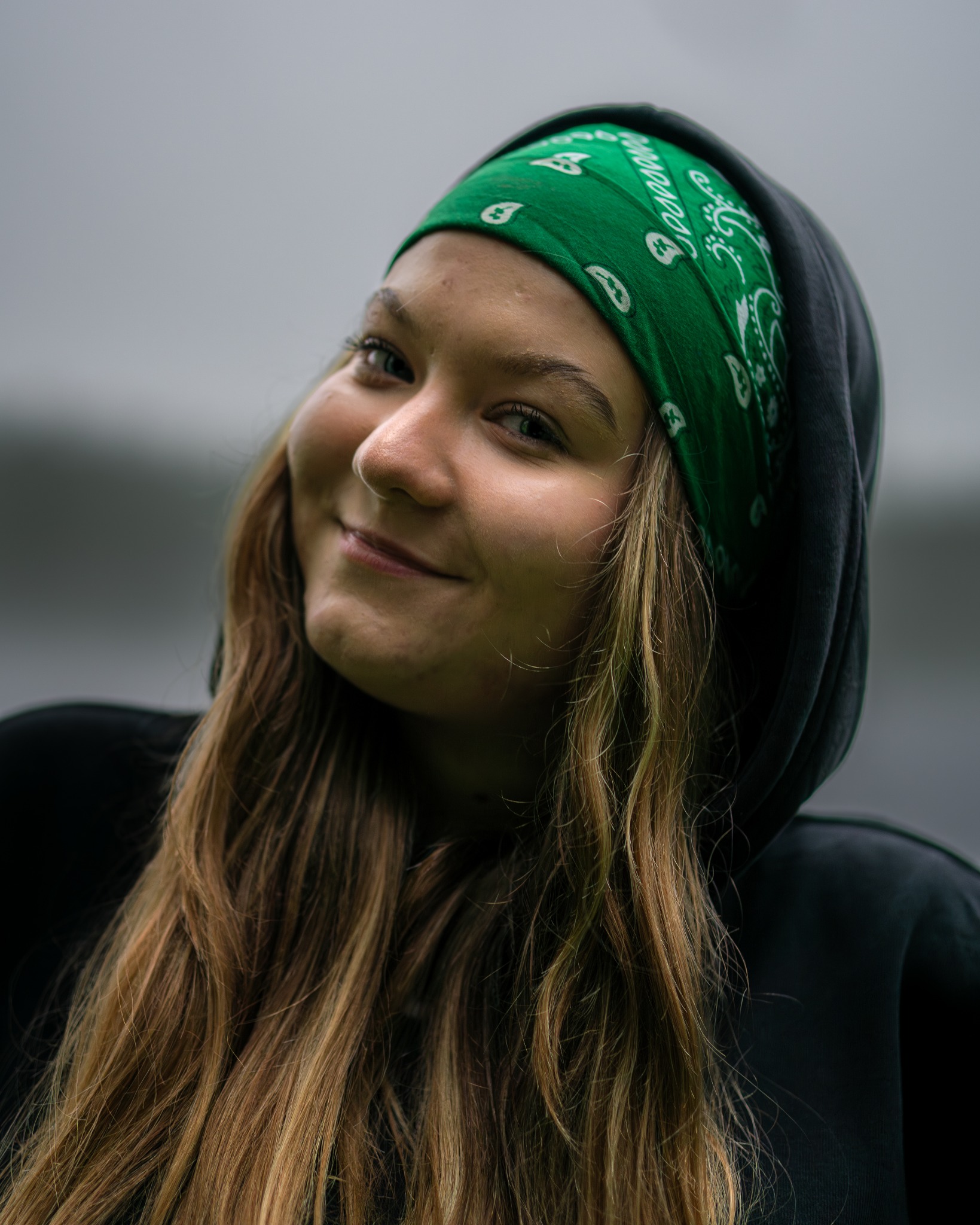 Portret uśmiechniętej młodej kobiety z zieloną bandaną na głowie, ubrana w czarną bluzę z kapturem, na rozmytym tle.