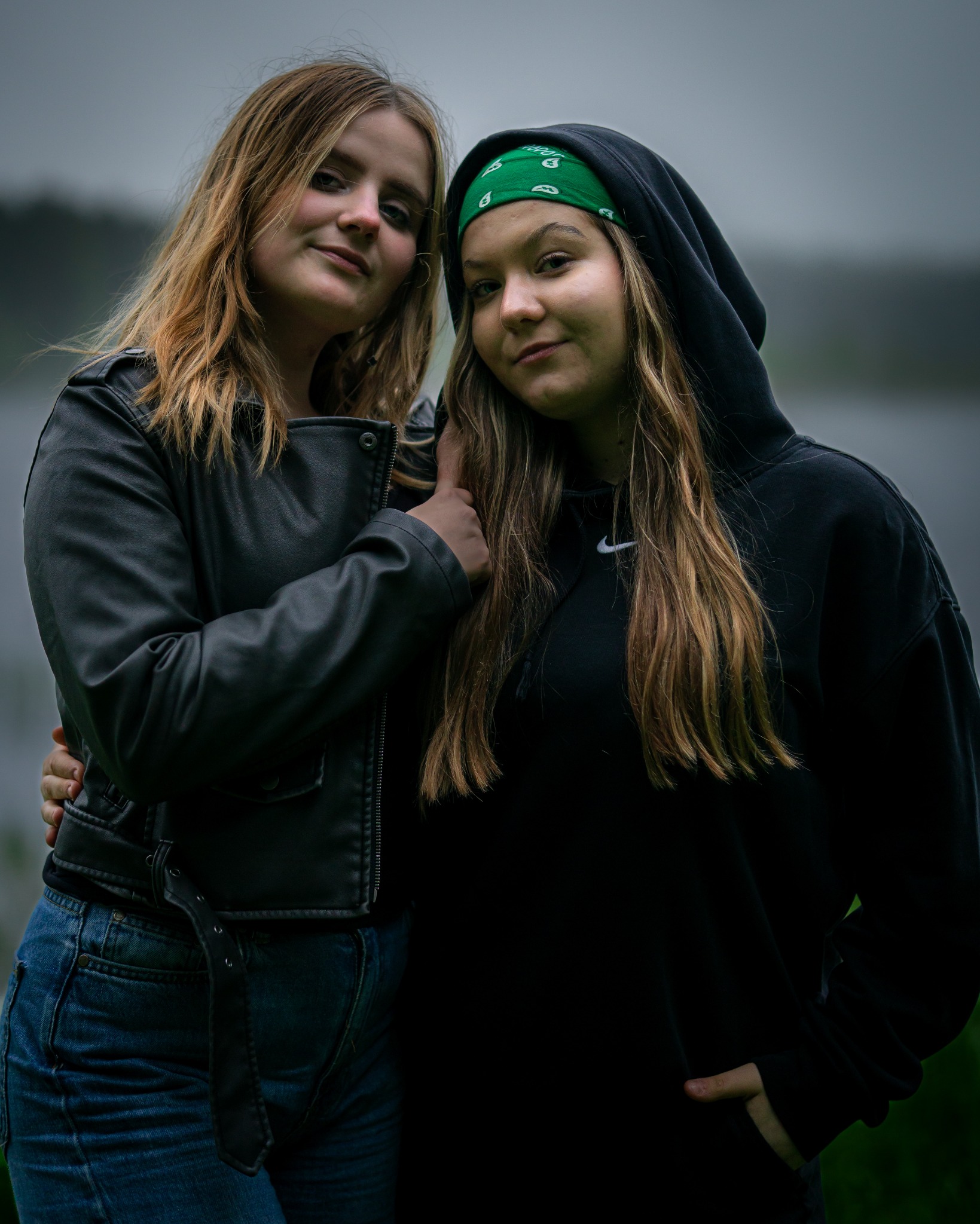 Dwie nastolatki przytulone, jedna w czarnej skórzanej kurtce i dżinsach, druga w czarnej bluzie z kapturem i zielonej bandanie, na rozmytym tle jeziora.