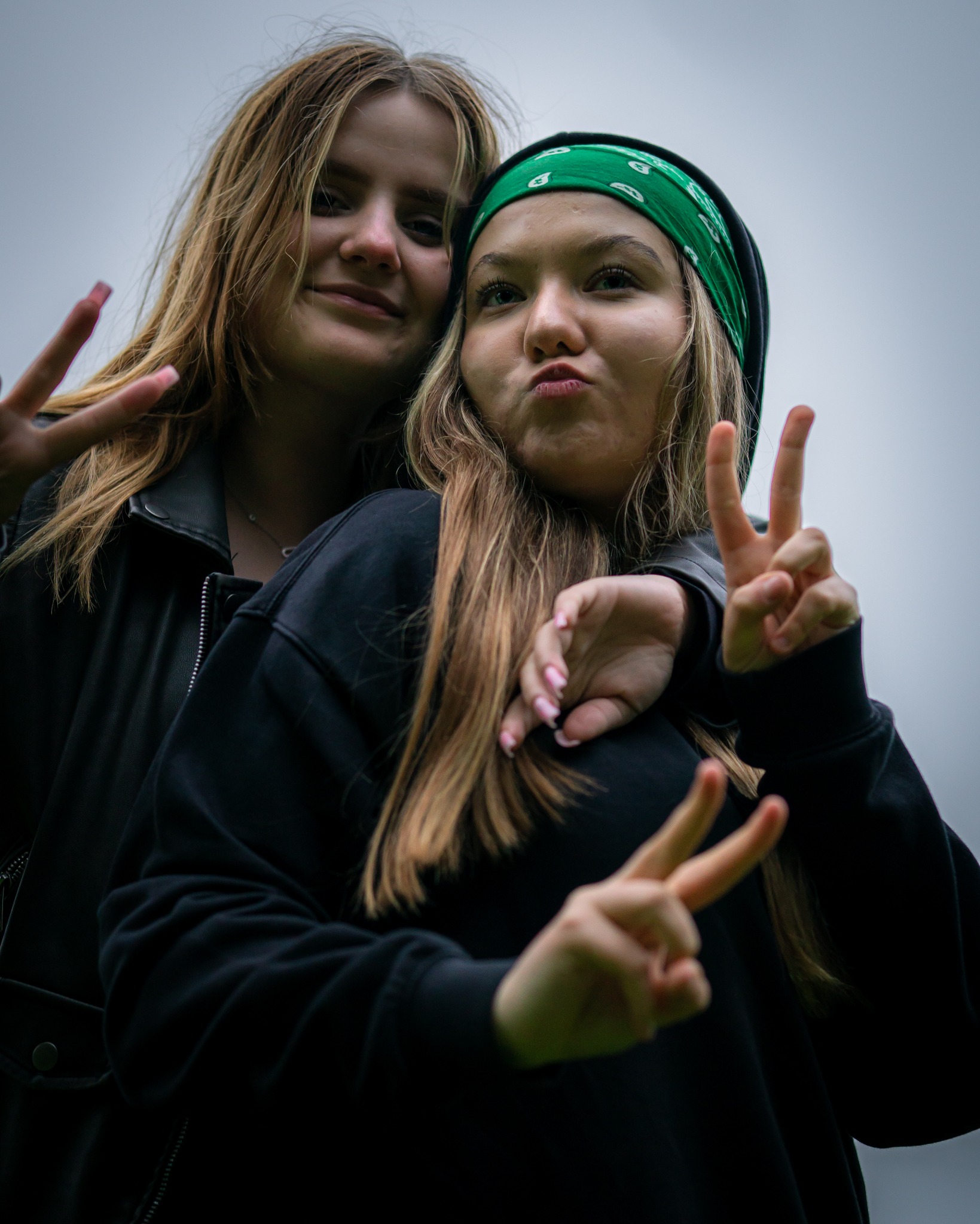 Dwie młode kobiety pozują, pokazując znak pokoju; jedna z zieloną bandaną, druga w skórzanej kurtce.