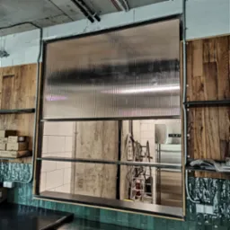 Przezroczysta, falista brama przemysłowa w pozycji otwartej, widok z wnętrza pomieszczenia, z widocznymi elementami instalacji elektrycznej i regałami po bokach.