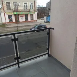 Szklana balustrada balkonowa z czarnymi profilami, widok na ulicę z zaparkowanym samochodem i budynkiem z balkonem.