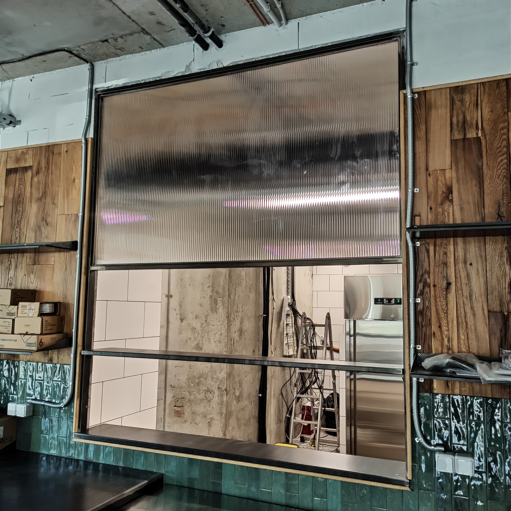 Przezroczysta, falista brama przemysłowa w pozycji otwartej, widok z wnętrza pomieszczenia, z widocznymi elementami instalacji elektrycznej i regałami po bokach.