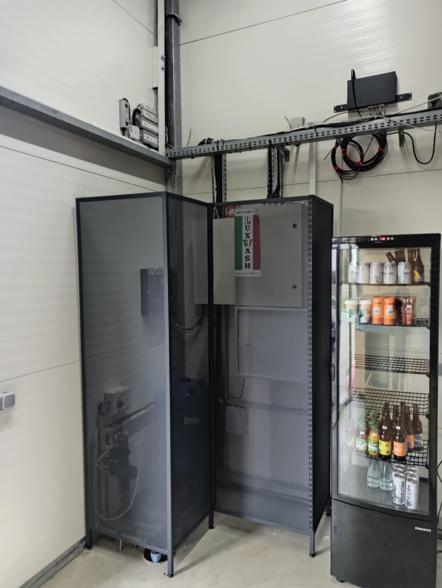 Metalowa obudowa urządzenia z perforowanej blachy, obok chłodziarka z napojami w hali produkcyjnej.