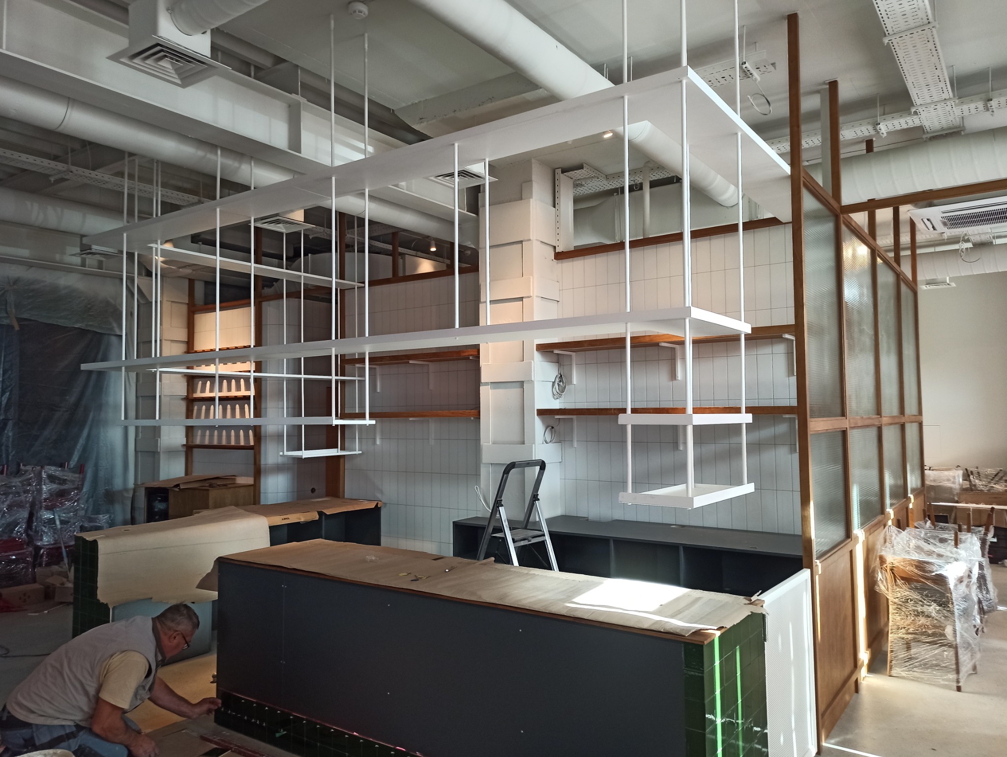 Wnętrze lokalu w trakcie wykańczania: montaż wiszących, metalowych konstrukcji regałowych w białym kolorze, widoczna drabina, częściowo zakryte meble i pracownik układający płytki.