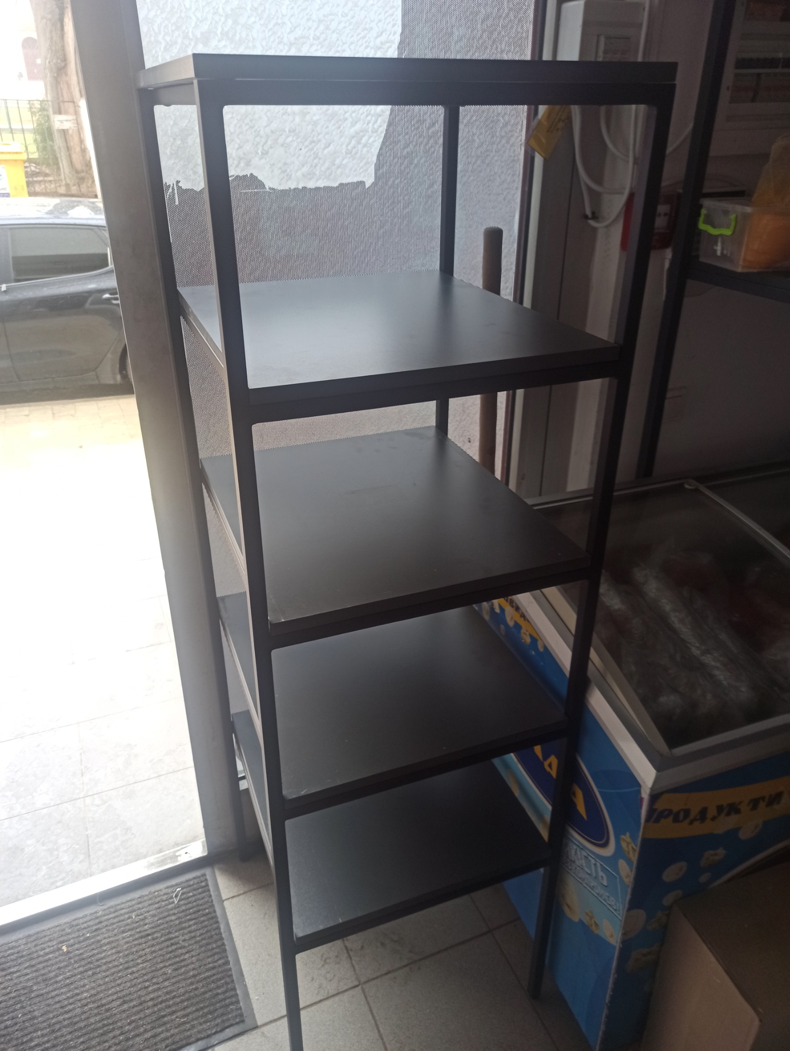 Metalowy regał z pięcioma półkami o surowym, minimalistycznym wyglądzie, ustawiony w pomieszczeniu z widocznym wejściem i chłodnicą w tle.