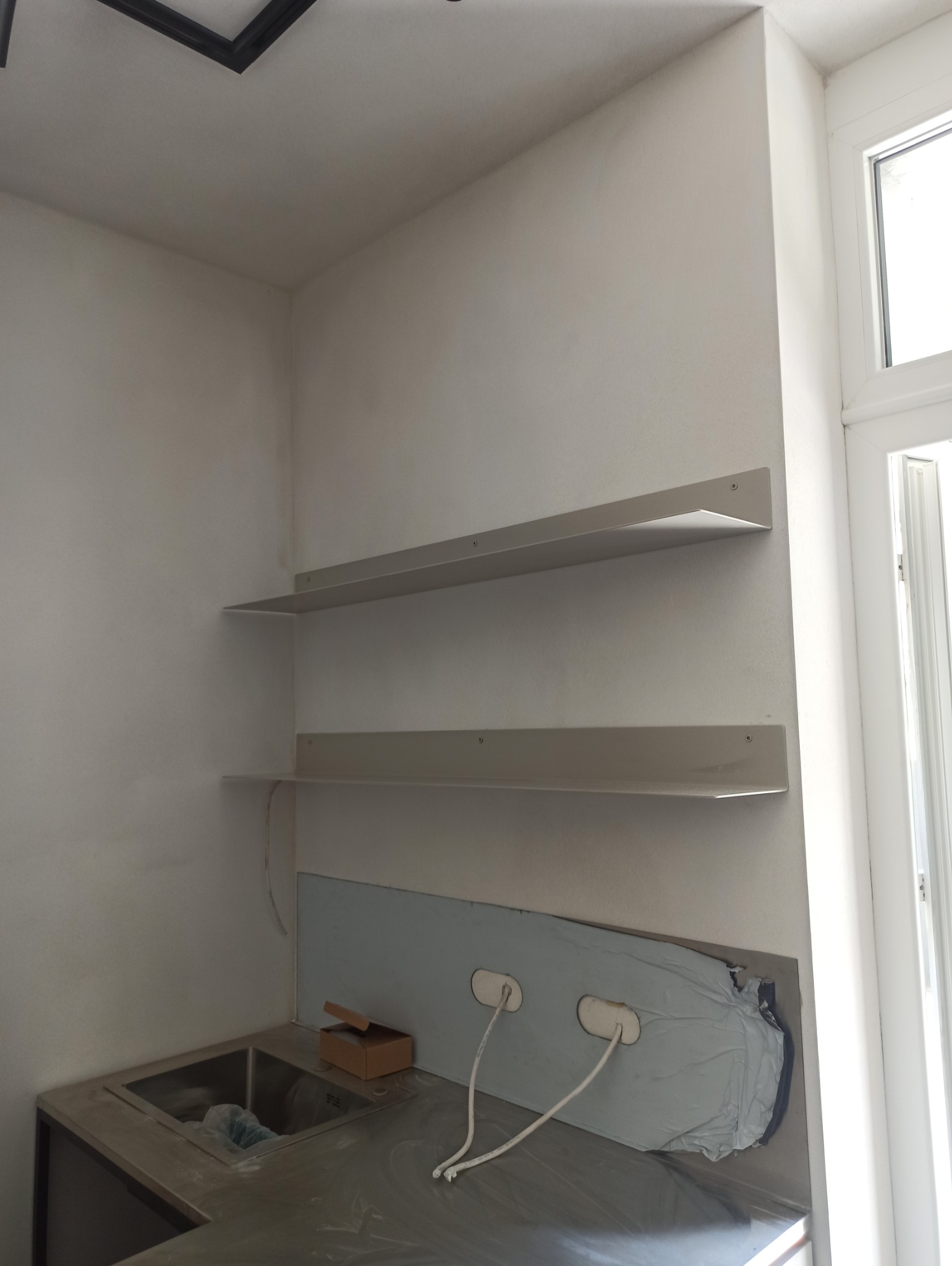 Dwie metalowe półki zamocowane na ścianie nad zlewem w trakcie remontu kuchni, widoczne kable i zabezpieczenia blatu.