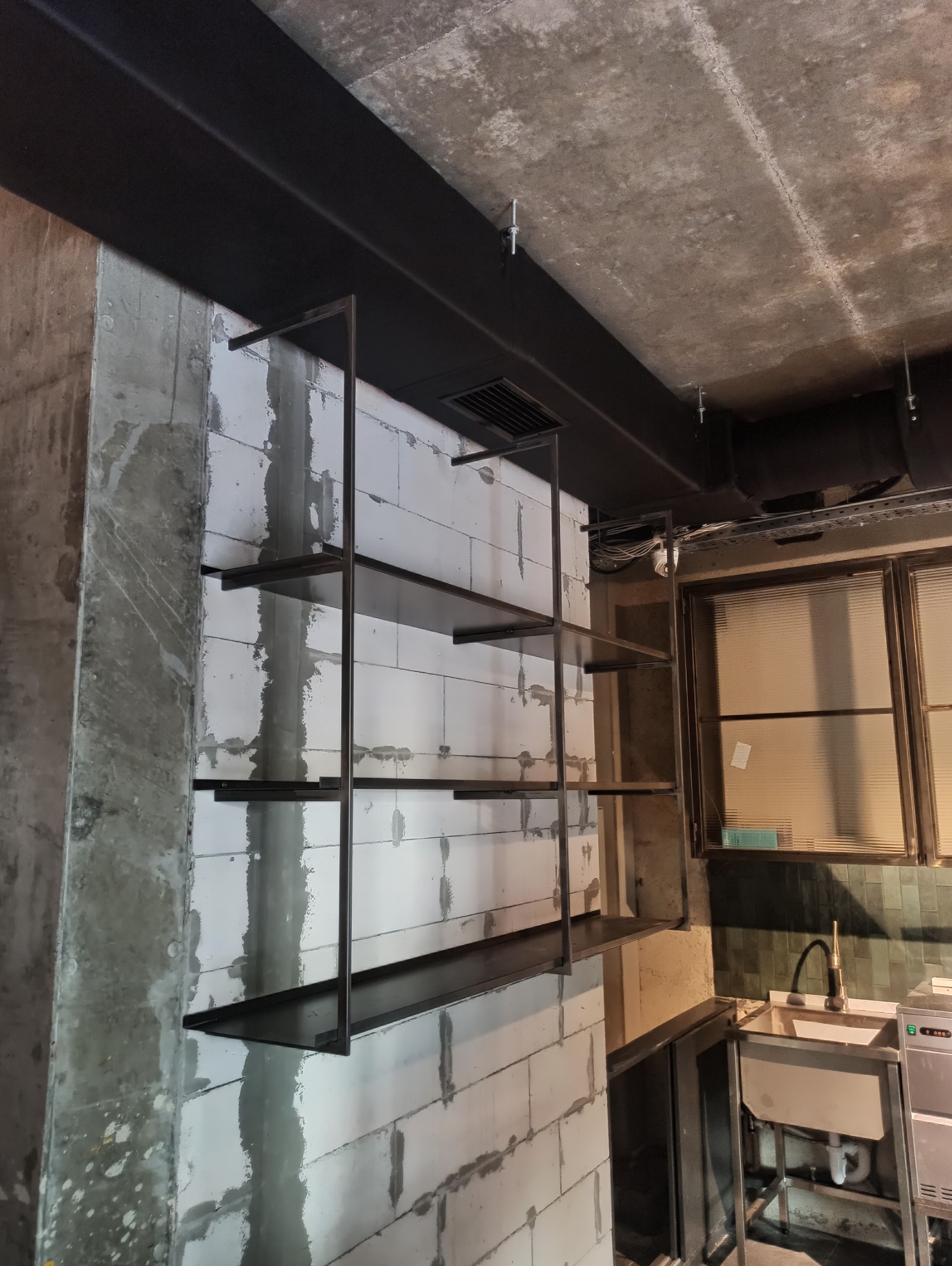 Metalowa konstrukcja regału z półkami, zamontowana na ścianie z betonu komórkowego w surowym wnętrzu z widoczną instalacją pod sufitem i fragmentem kuchni.