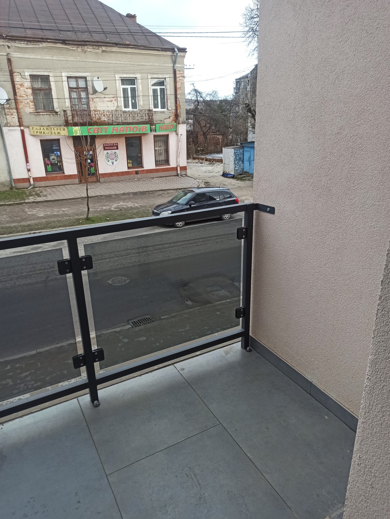 Szklana balustrada balkonowa z czarnymi profilami, widok na ulicę z zaparkowanym samochodem i budynkiem z balkonem.