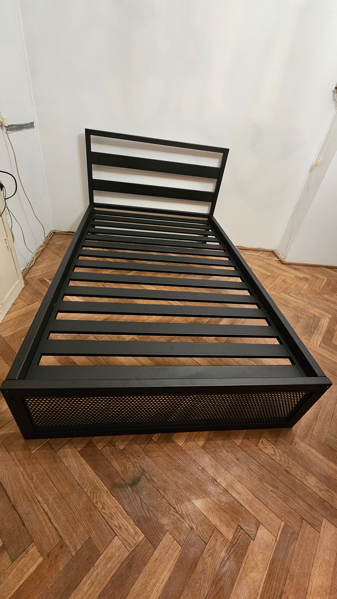 Metalowa rama łóżka z kratką u dołu, na parkiecie ułożonym w jodełkę, w pokoju z białymi ścianami.