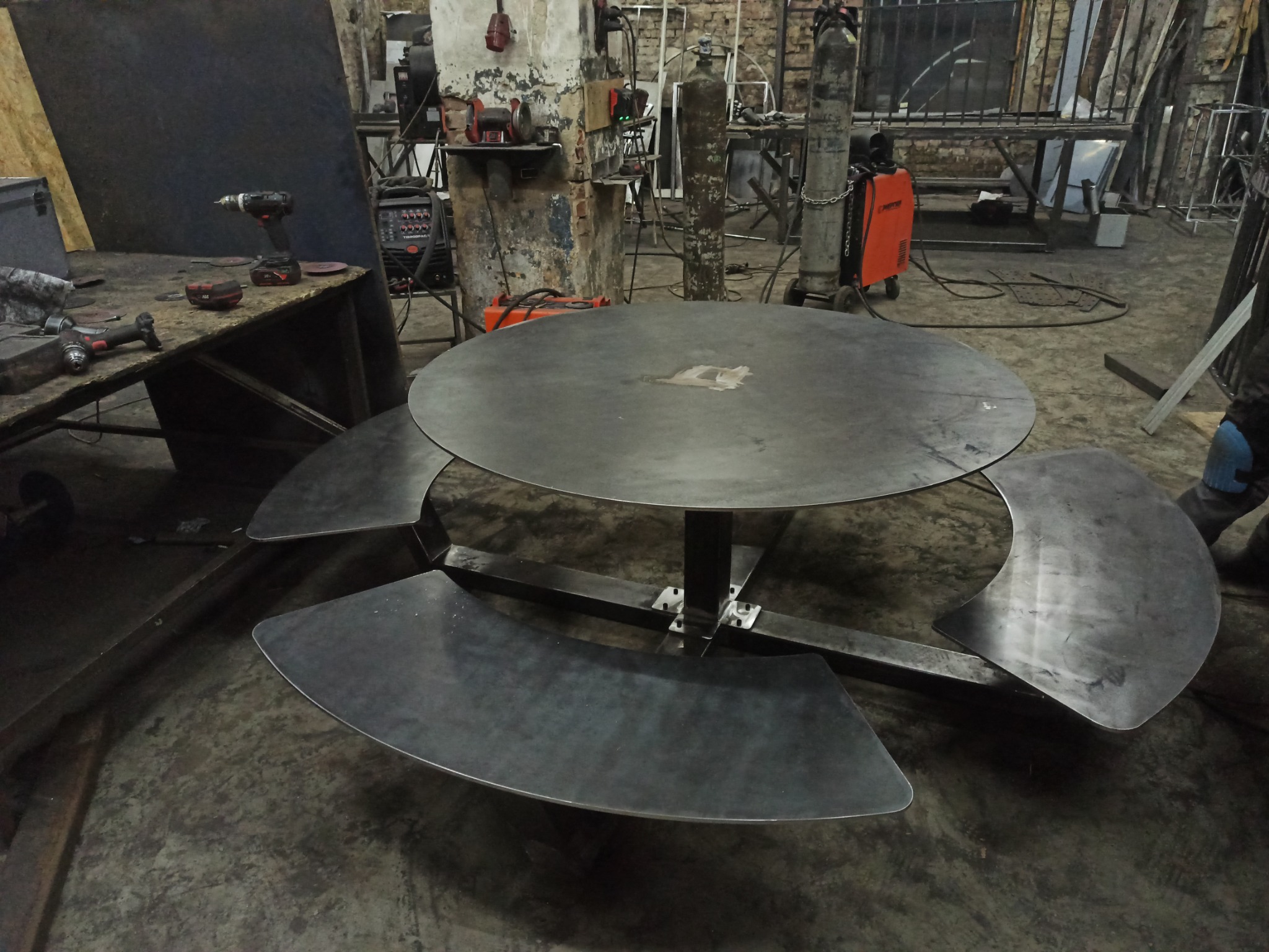 Okrągły, metalowy stół z trzema półokrągłymi, metalowymi siedziskami w warsztacie, widoczne spawarki i narzędzia w tle.