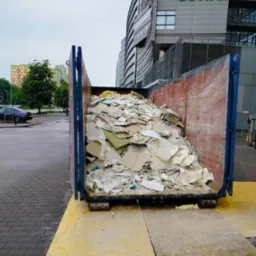 Kontener wypełniony gruzem budowlanym, głównie fragmentami płyt gipsowo-kartonowych, ustawiony na żółtej płycie OSB w miejskiej scenerii z budynkami biurowymi w tle.