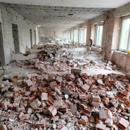 EKO TRANS - Wnętrze budynku w trakcie rozbiórki, gruz ceglany i betonowy na podłodze, widoczne ściany z odsłoniętą cegłą i okna z białymi ramami, w tle sylwetki pracowników w odblaskowych kamizelkach.