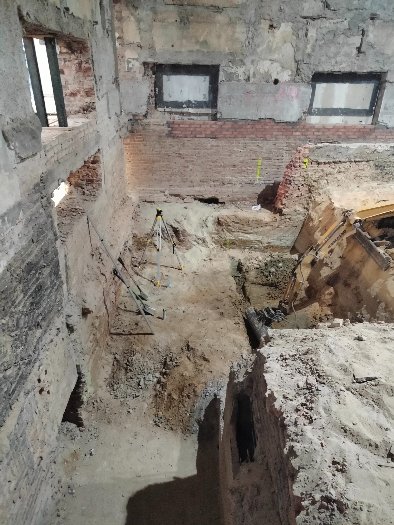 Wnętrze budynku w trakcie prac ziemnych: wykop z żółtą koparką Komatsu, niwelator geodezyjny, łopaty i resztki ceglanych ścian z oknami.