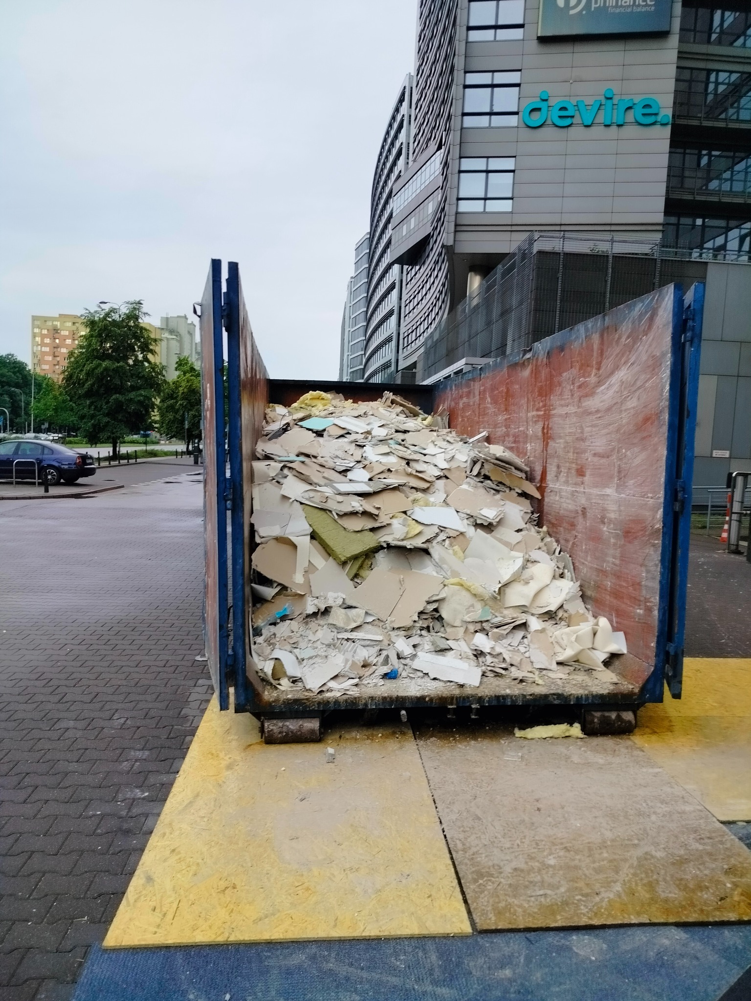 Kontener wypełniony gruzem budowlanym, głównie fragmentami płyt gipsowo-kartonowych, ustawiony na żółtej płycie OSB w miejskiej scenerii z budynkami biurowymi w tle.