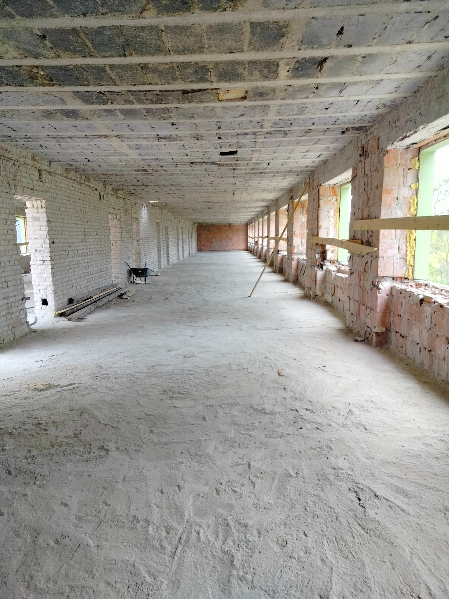 Długie, puste pomieszczenie w trakcie remontu; surowe ściany z cegły, betonowy sufit, prowizoryczne zabezpieczenia okien, w oddali taczka budowlana.