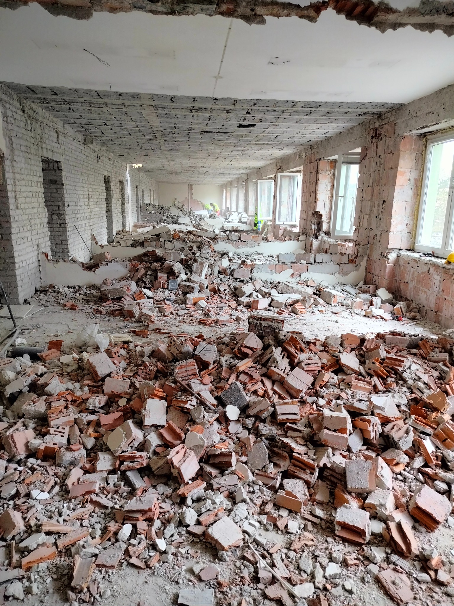 Wnętrze budynku w trakcie rozbiórki, gruz ceglany i betonowy na podłodze, widoczne ściany z odsłoniętą cegłą i okna z białymi ramami, w tle sylwetki pracowników w odblaskowych kamizelkach.