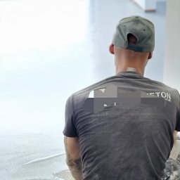 DJ art floor & walls - Pracownik w trakcie aplikacji mikrocementu na podłogę w jasnym pomieszczeniu, widok z tyłu, widoczny fragment logo firmy na koszulce.