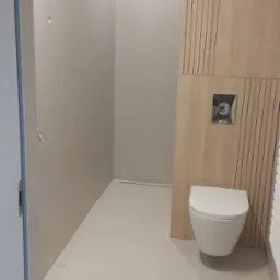 Minimalistyczna łazienka z mikrocementową podłogą, drewnianą boazerią i podwieszaną toaletą. Widoczny odpływ liniowy w podłodze.