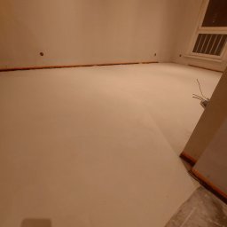 DJ art floor & walls - Jasna, gładka posadzka mikrocementowa w trakcie realizacji, zabezpieczona pomarańczową taśmą na styku ze ścianą, widoczne kable elektryczne wystające ze ściany.