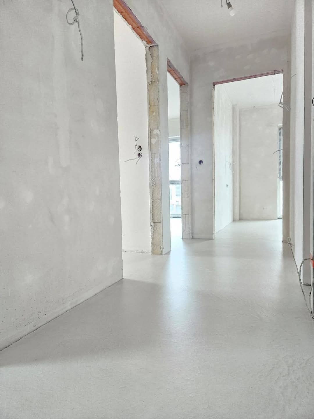 Wnętrze w trakcie remontu z gładką, jasnoszarą posadzką, surowe ściany i framugi drzwiowe, widoczne przewody elektryczne.