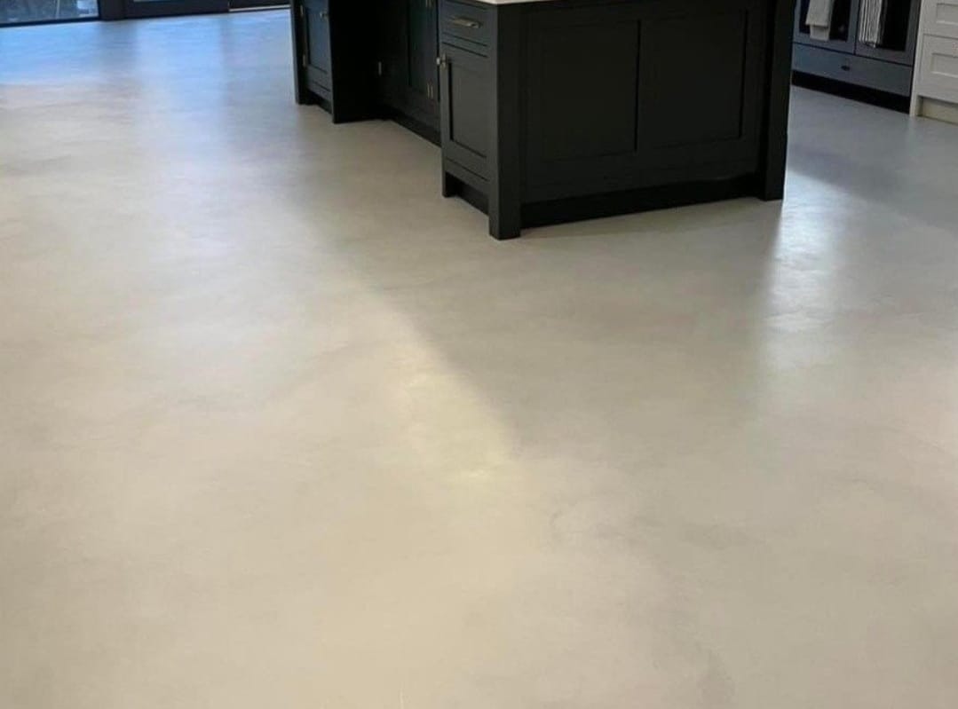 Jasnoszara, gładka posadzka w kuchni z ciemną wyspą kuchenną. Widoczne delikatne cieniowanie na powierzchni podłogi.