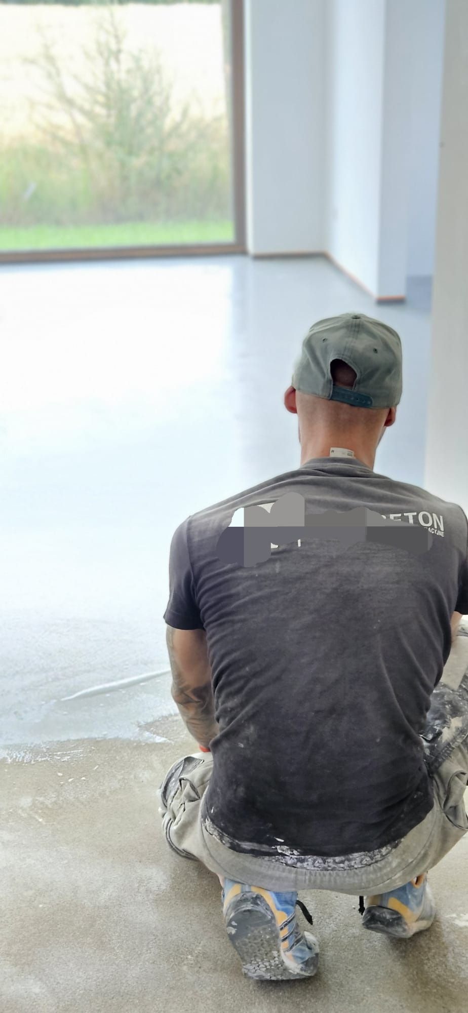 Pracownik w trakcie aplikacji mikrocementu na podłogę w jasnym pomieszczeniu, widok z tyłu, widoczny fragment logo firmy na koszulce.