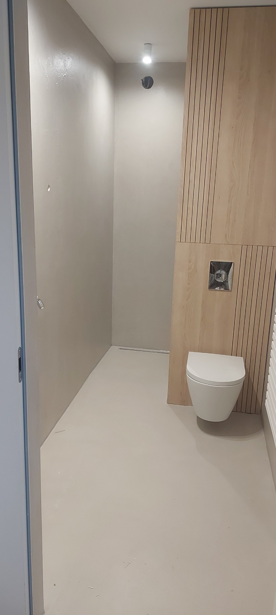 Minimalistyczna łazienka z mikrocementową podłogą, drewnianą boazerią i podwieszaną toaletą. Widoczny odpływ liniowy w podłodze.