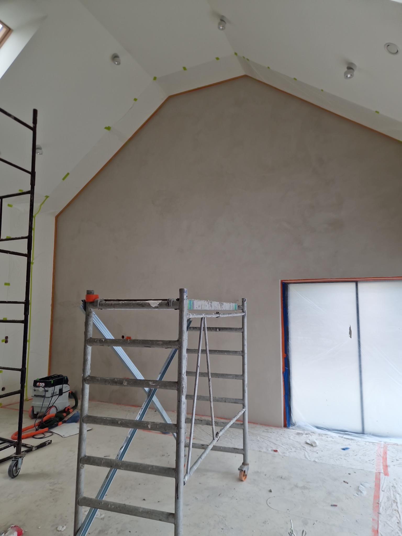 Ściana pokryta mikrocementem w trakcie aplikacji, widoczne rusztowania, taśmy malarskie zabezpieczające krawędzie i folia ochronna na oknie.