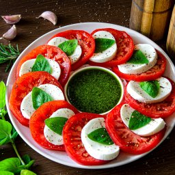 Mikołaj Dyraga - Caprese sałatka z pomidorów, mozzarelli i pesto na białym talerzu, udekorowana bazylią i czosnkiem, na ciemnym drewnianym stole.