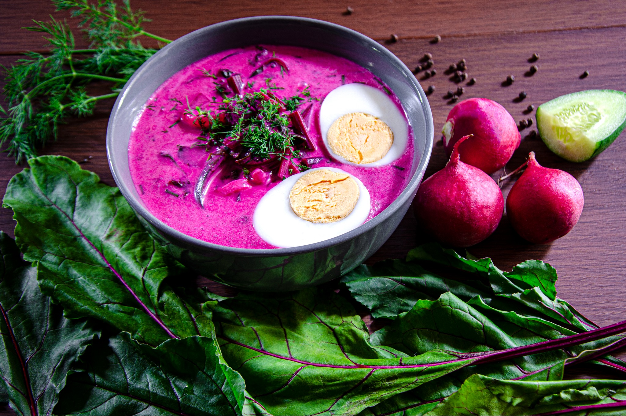 Chłodnik litewski w szarej misce, udekorowany koperkiem i jajkiem na twardo, obok rzodkiewki, ogórek i liście buraka na drewnianym stole.