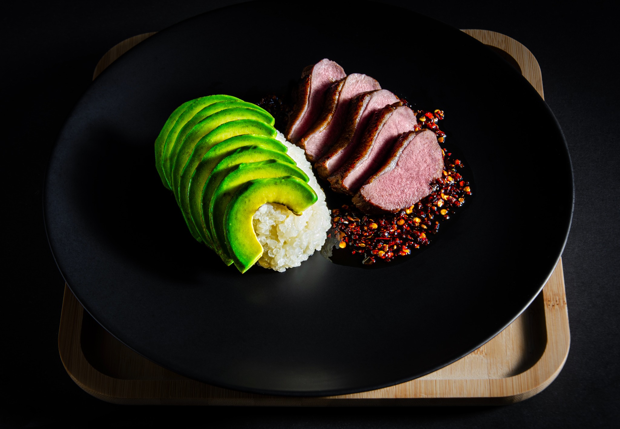 Elegancko podane danie: plastry kaczki z sosem chili, awokado ułożone wachlarzowo i porcja ryżu na czarnym talerzu, całość na drewnianej podkładce.