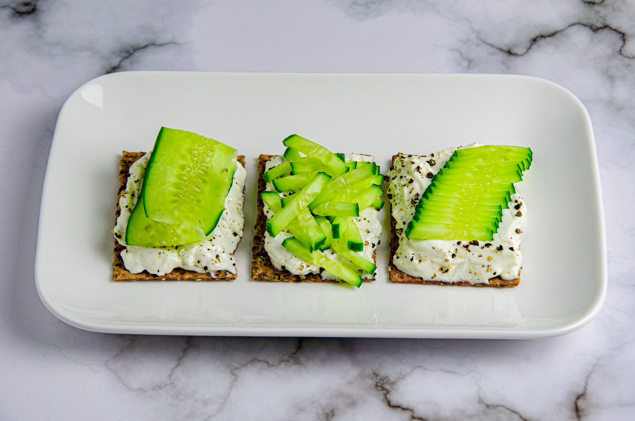 Trzy kanapki na kwadratowych krakersach z kremowym serem i różnymi sposobami ułożonymi plasterkami ogórka na białym talerzu na marmurowym tle.