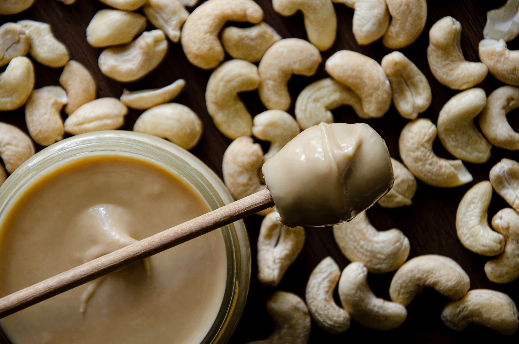 Słoik kremu z orzechów nerkowca, drewniana łyżeczka z kremem i rozsypane orzechy nerkowca na ciemnym drewnianym tle, ujęcie z góry.