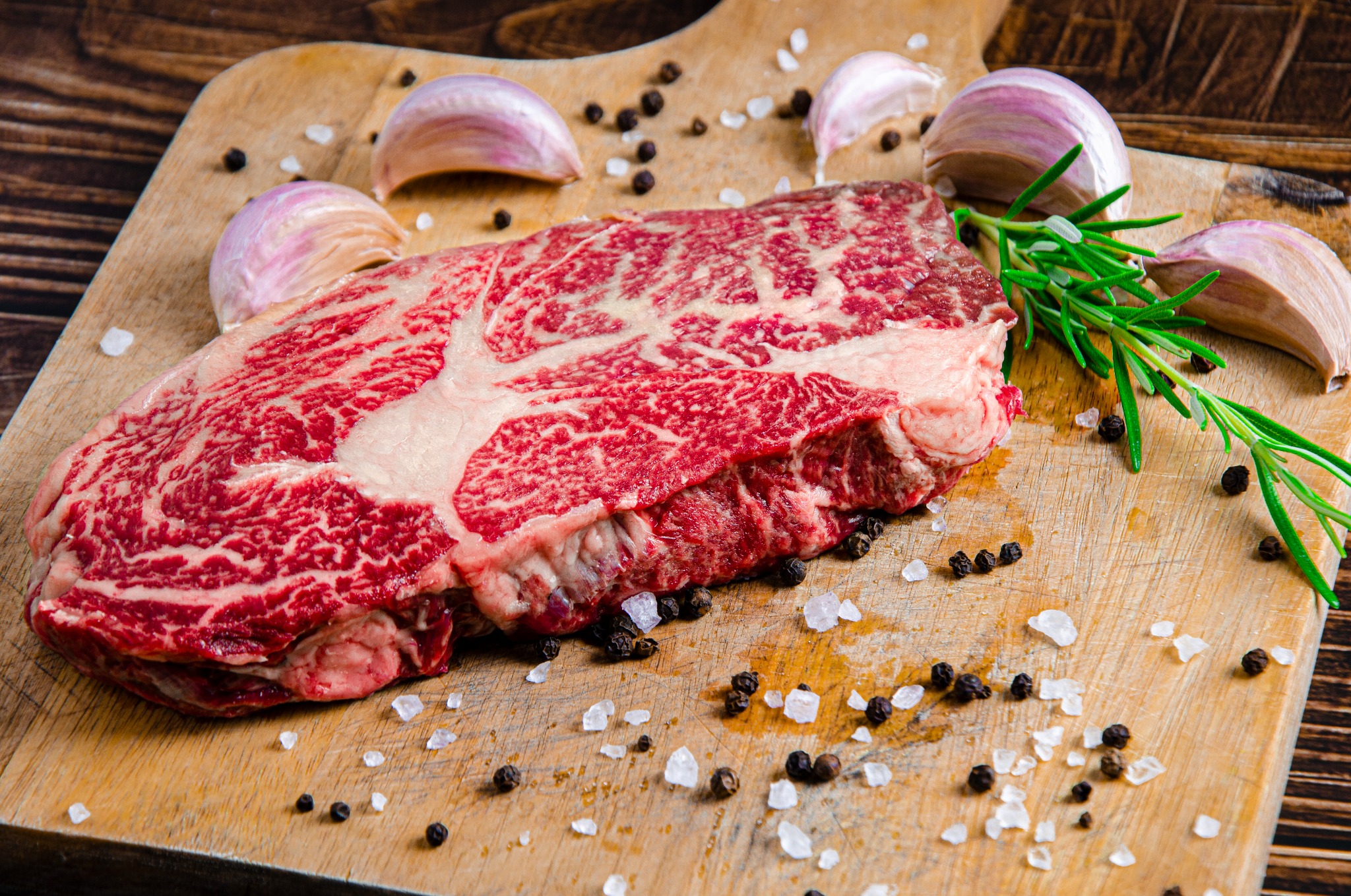 Surowy stek wołowy z marmurkowym wzorem, czosnkiem, rozmarynem, solą i pieprzem na drewnianej desce do krojenia.