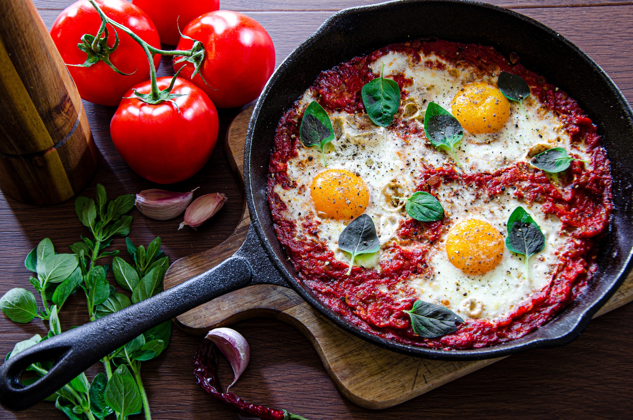 Shakshuka w żeliwnej patelni na drewnianej desce, ozdobiona świeżą bazylią, z pomidorami i czosnkiem w tle.