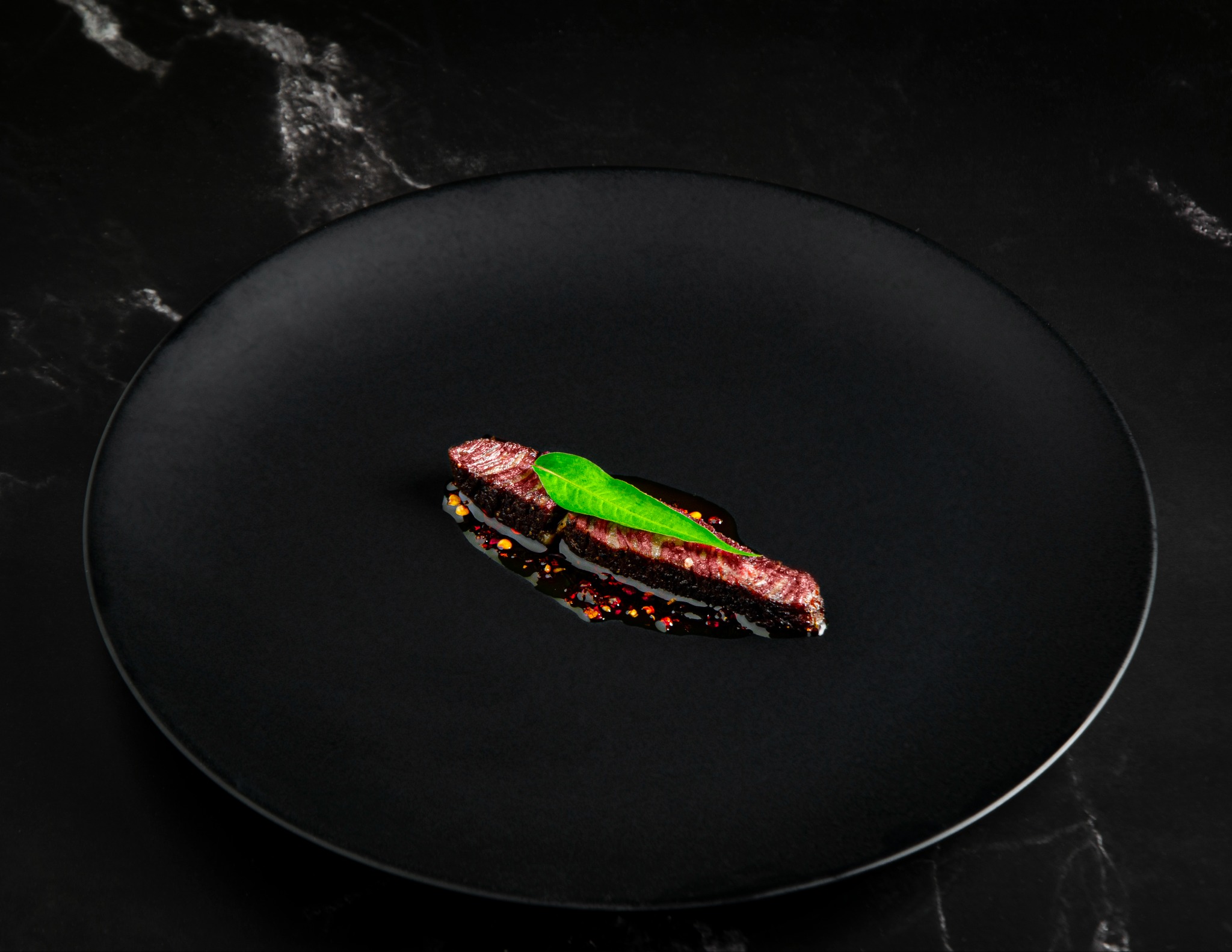 Fotografia produktowa: Pojedynczy plasterek krwistego steka z zielonym listkiem na czarnym talerzu na tle marmurowego blatu.