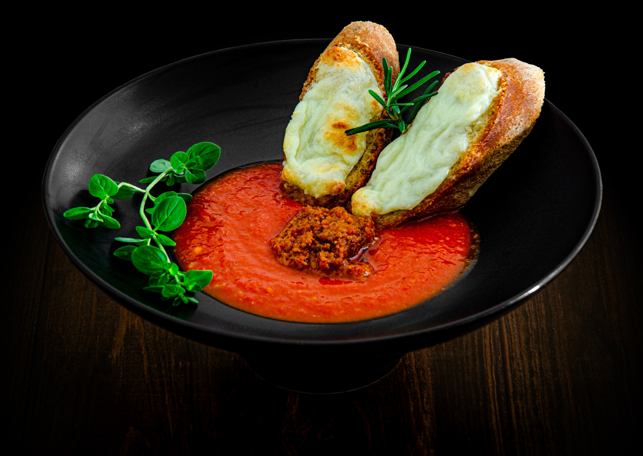 Dwa grzanki z zapieczonym serem, podane na talerzu z sosem pomidorowym i pesto, udekorowane gałązką rozmarynu i oregano, na ciemnym drewnianym tle.