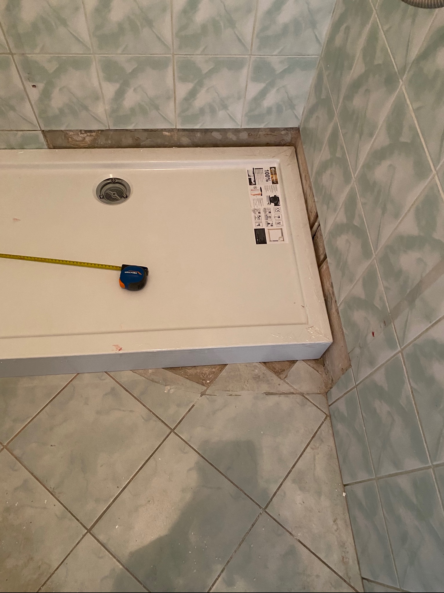 Biały brodzik prysznicowy z odpływem, częściowo owinięty folią ochronną, mierzony taśmą mierniczą Dexter, w rogu łazienki z zielonymi płytkami, widoczne ślady po demontażu.