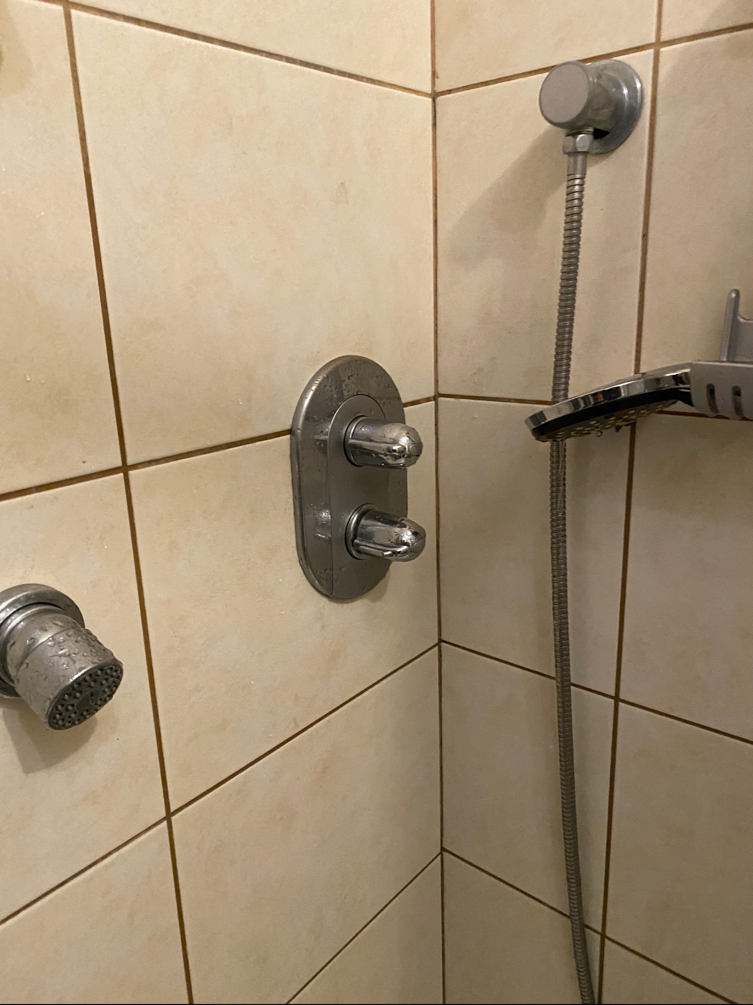 Szczegółowy widok baterii prysznicowej z chromowanymi elementami na beżowych, kwadratowych płytkach ceramicznych. Widoczne ślady wody na baterii i słuchawce prysznicowej.