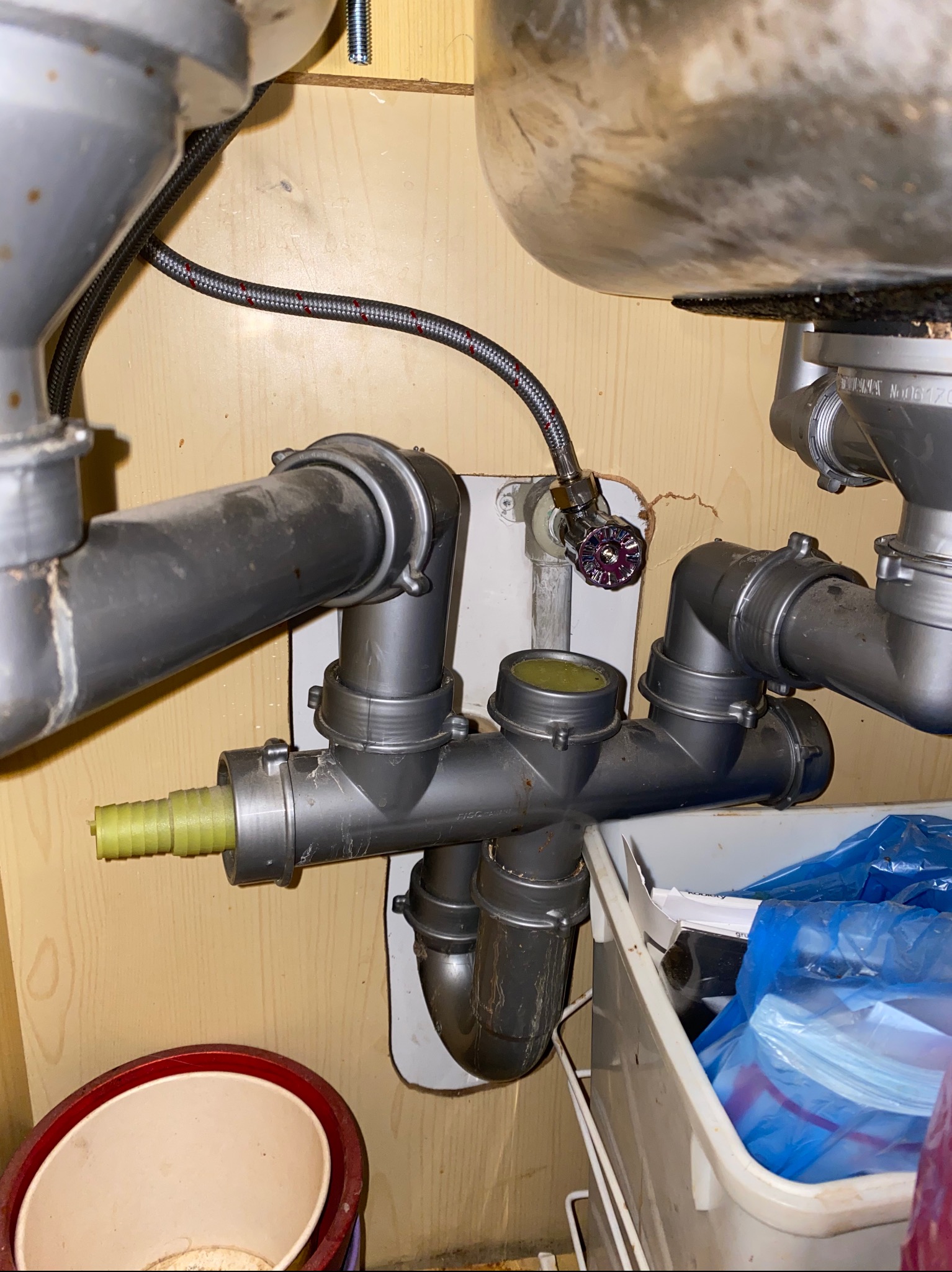 Szare rury kanalizacyjne pod zlewem, widoczny zawór wody z bordową rączką, żółta końcówka węża, wnętrze szafki z elementami instalacji.